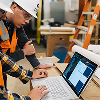 Two construction workers in safety gear and hard hats focus on a laptop displaying plans, suggesting a collaborative and focused work environment.