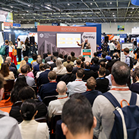 A speaker addresses a packed audience at a tech conference. Attendees are focused on a presentation screen, creating a lively and engaged atmosphere.