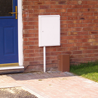 A white utility box mounted on a red brick wall next to a vibrant blue door. The scene includes a paved walkway and a small patch of grass.