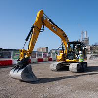 A yellow excavator on a construction site with two workers beside it under a clear blue sky. Nearby barriers and distant buildings are visible.